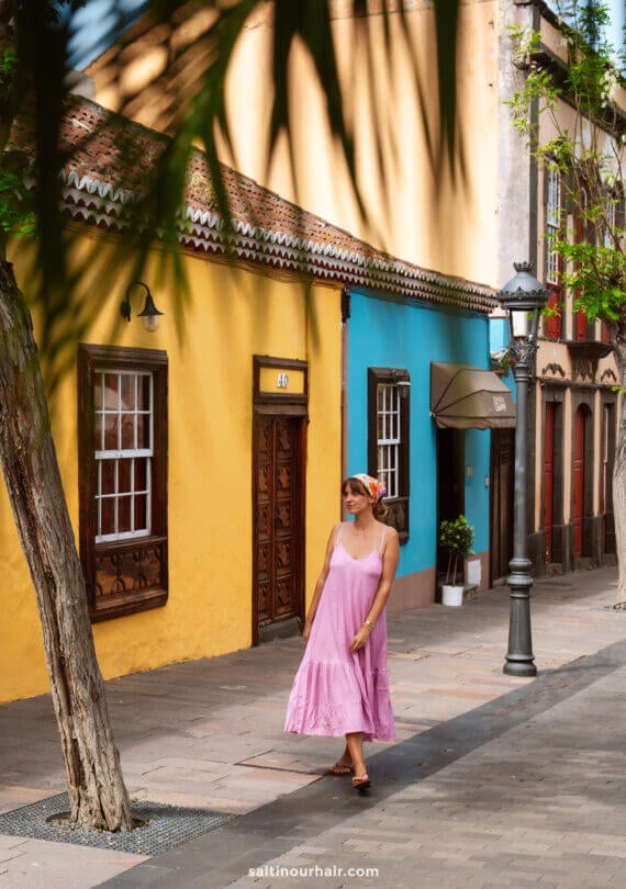 A woman in a pink dress and headscarf strolls along a sunlit street in La Palma, with colorful yellow and blue buildings, trees, and street lamps—perfect for any Canary Islands travel guide.
