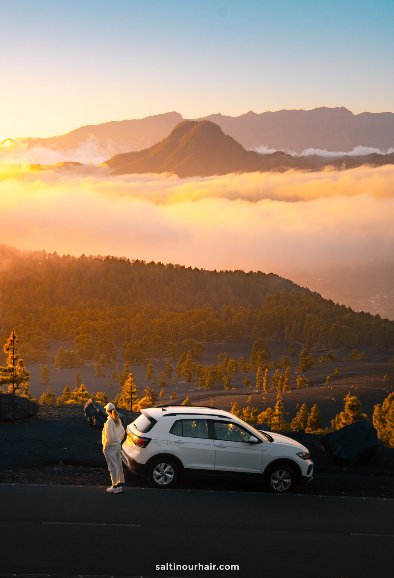 A person stands next to a white SUV parked on a mountain road at sunrise in La Palma, overlooking a valley filled with clouds and surrounded by forested hills in the Canary Islands.