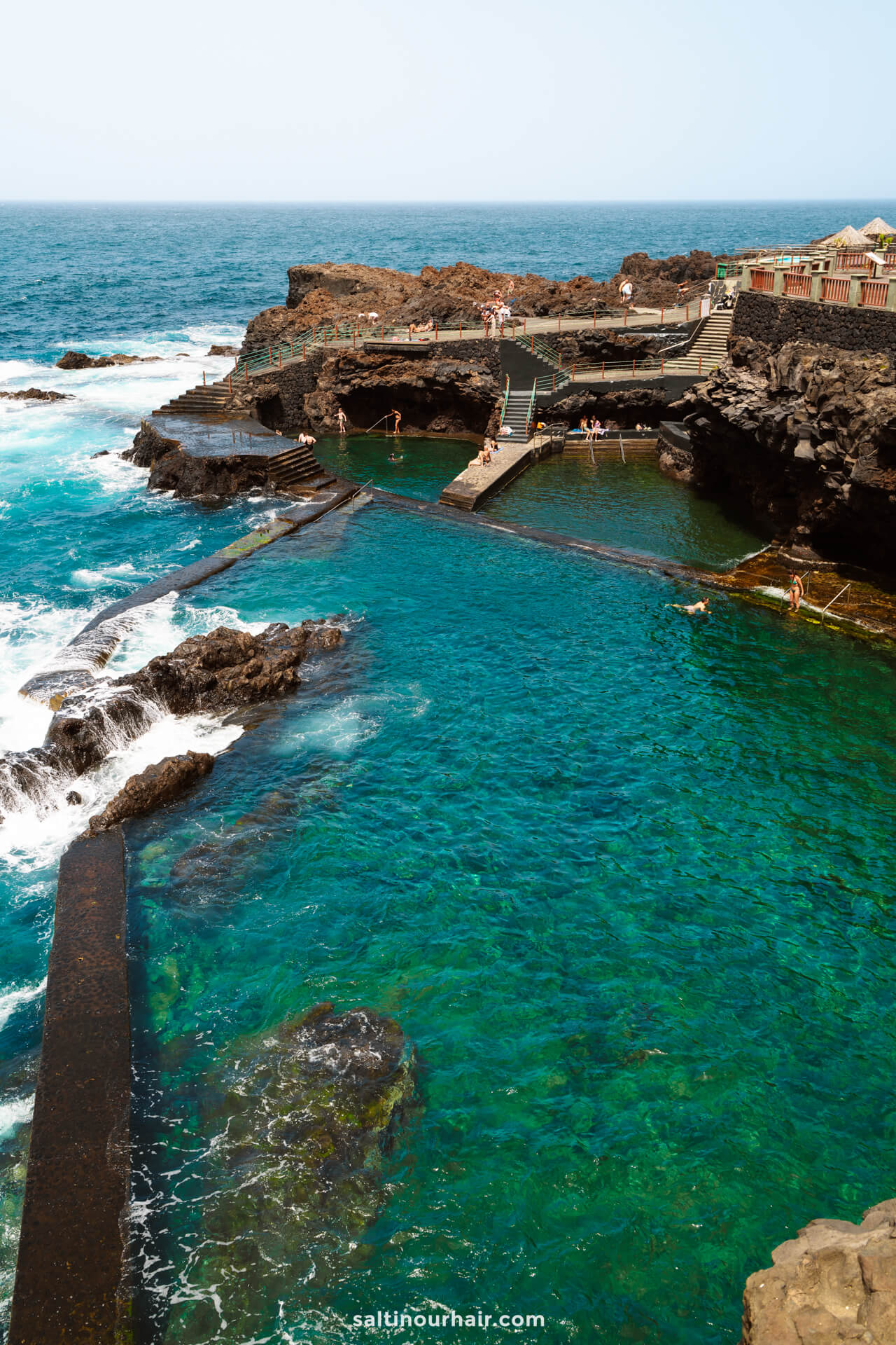 Natural volcanic rock pools with clear turquoise water, surrounded by rocky cliffs and people swimming, near the ocean under a clear sky—La Palma’s hidden gem in the Canary Islands, perfect for your next travel guide adventure.