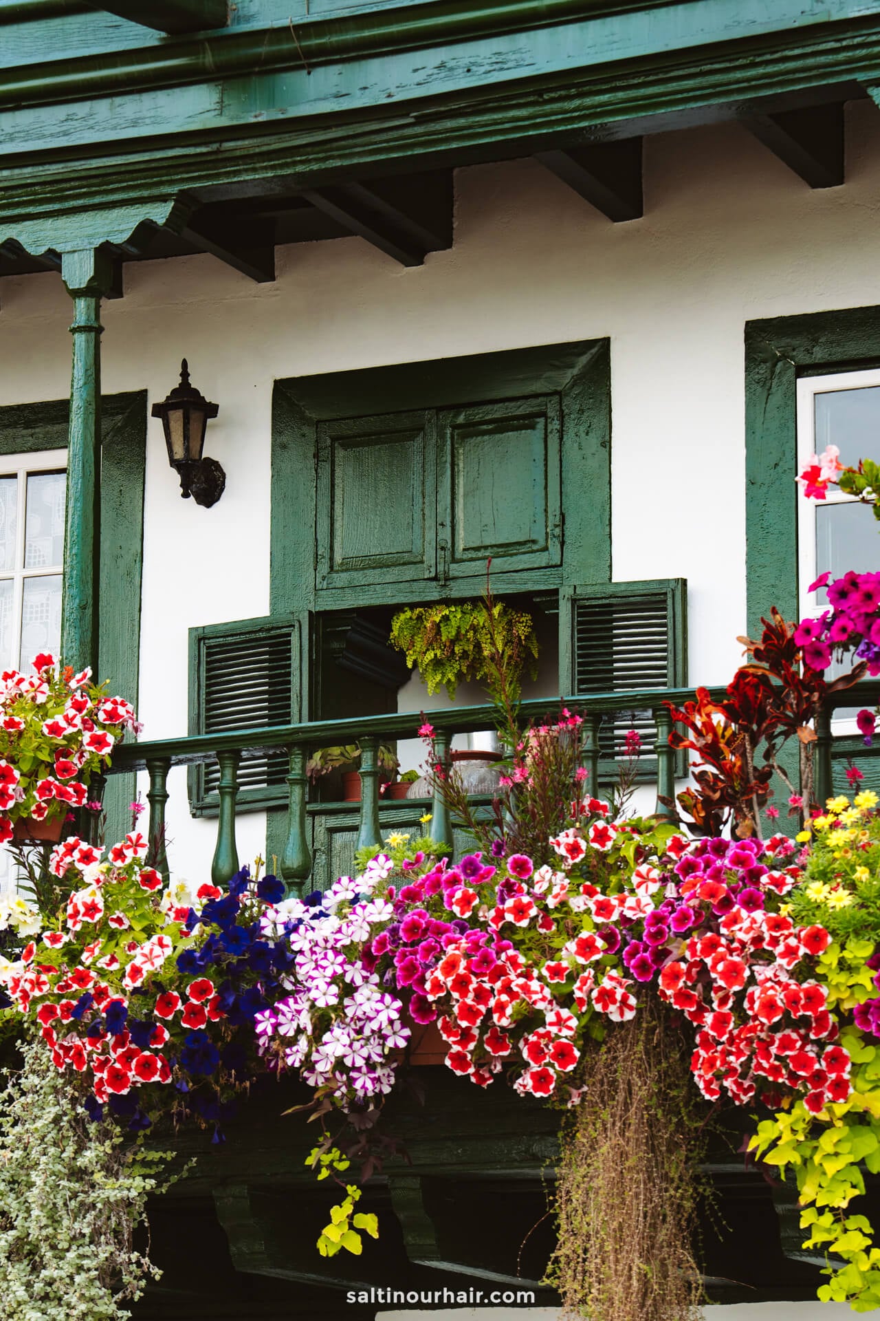A green wooden balcony with a half-closed window, decorated with a variety of colorful flowers, attached to a white building—typical charm found in La Palma, Canary Islands.