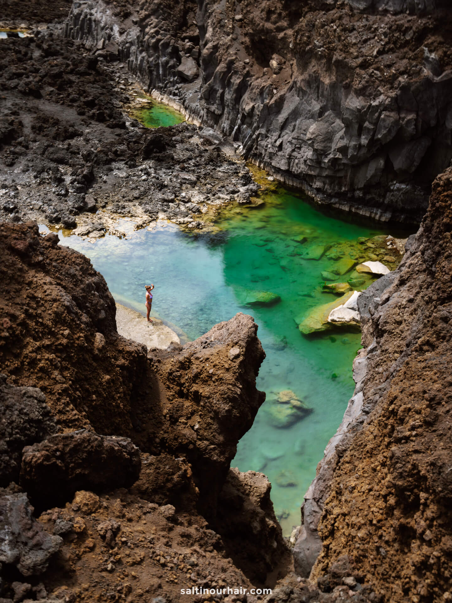 A person stands on rocky terrain beside a clear, turquoise stream surrounded by jagged, dark cliffs—a hidden gem in La Palma, one of the Canary Islands.