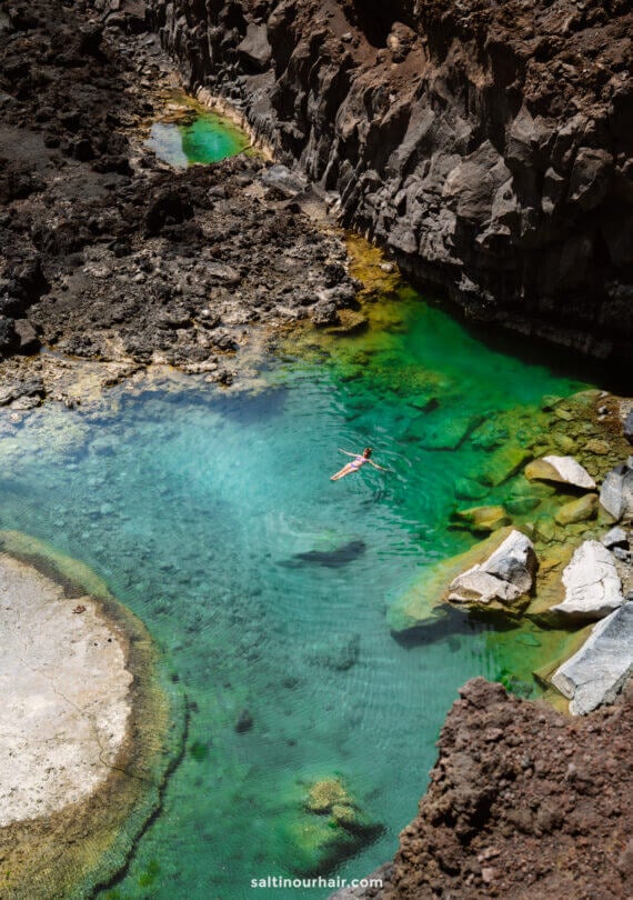 A person floats in a clear turquoise pool on La Palma, surrounded by rocky cliffs and dark volcanic terrain—a hidden gem of the Canary Islands featured in any top travel guide.