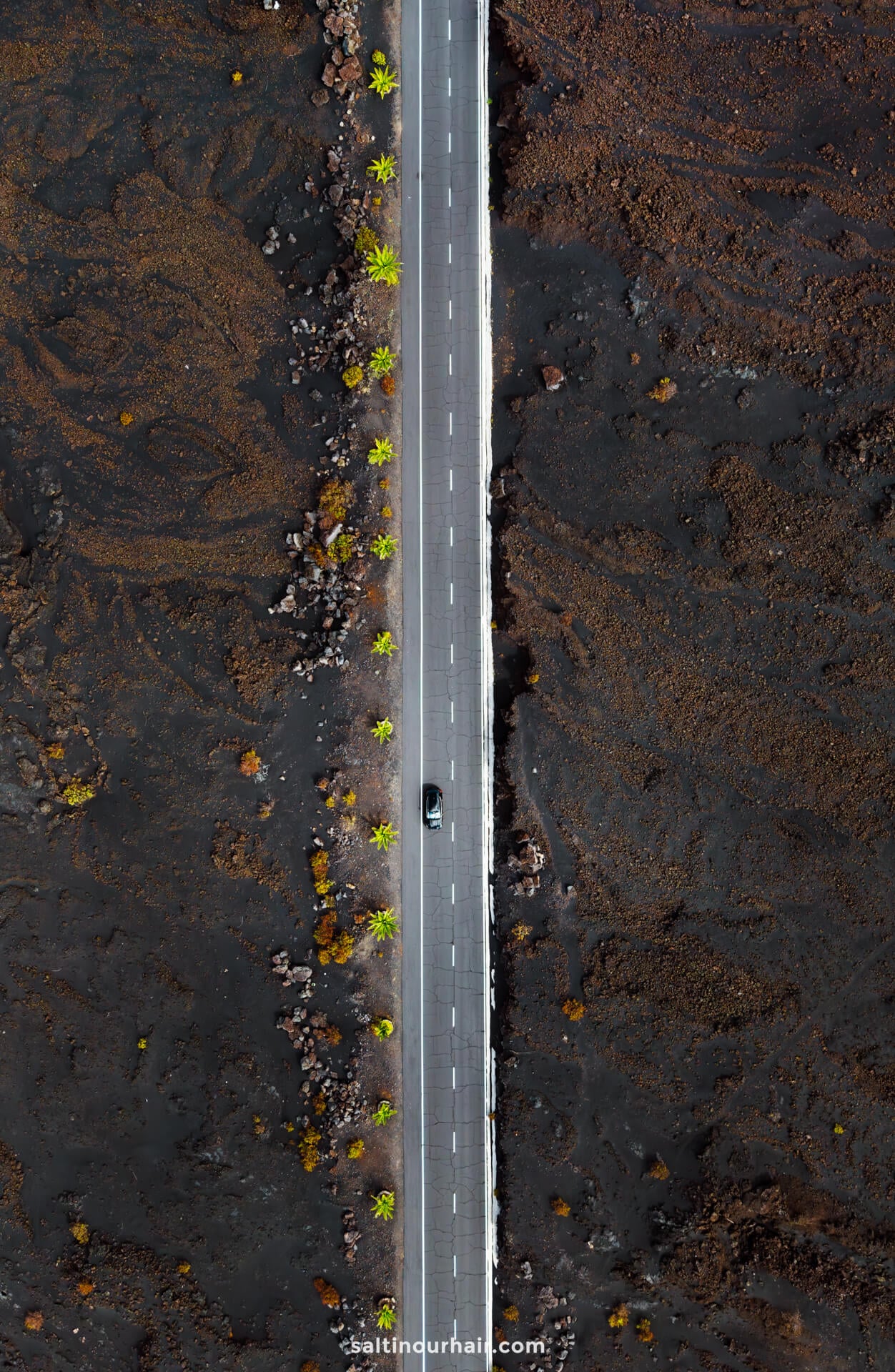 Aerial view of a straight road cutting through a barren, dark landscape on La Palma, Canary Islands, with small green trees lining both sides and a single car driving along it—perfect inspiration for your next travel guide.