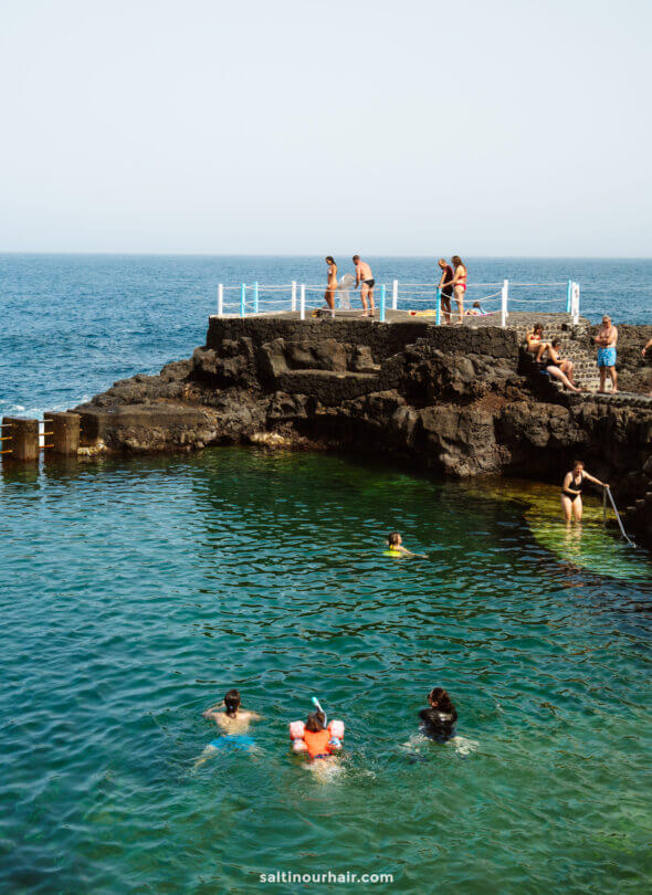 People swim and relax in a natural rock pool by the ocean on La Palma, with some climbing and jumping from the rocks; calm, clear water and a blue sky create a perfect scene from a Canary Islands travel guide.
