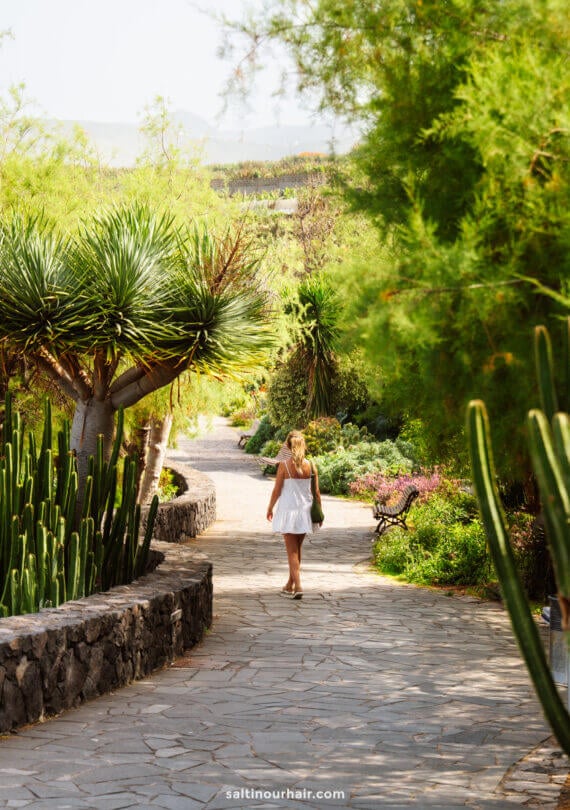A woman in a white dress strolls down a stone path in a lush, sunlit botanical garden on La Palma, surrounded by cacti and green plants—a scene perfect for any Canary Islands travel guide.