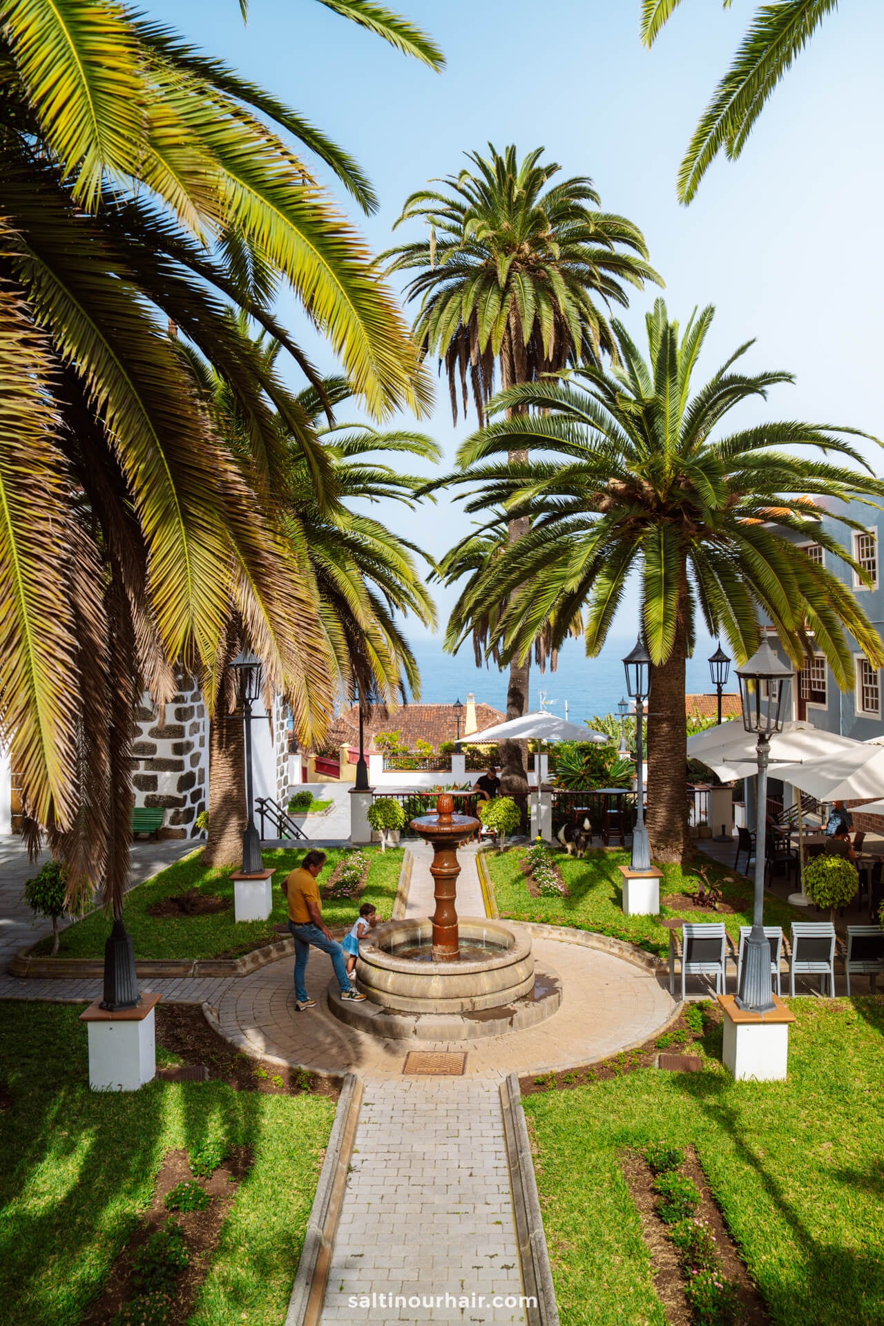 A man walks with a child near a circular fountain surrounded by palm trees and colonial-style buildings on a sunny day in La Palma, Canary Islands—a perfect snapshot for any travel guide.