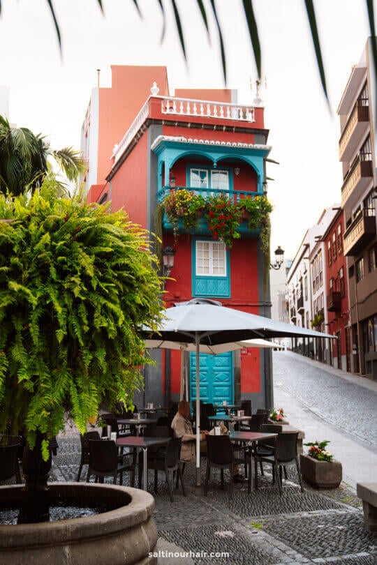 A red building with blue balconies and doors sits at a street corner; cafe tables with an umbrella and plants are in the foreground, echoing the vibrant charm of La Palma in the Canary Islands—perfect for your next travel guide inspiration.