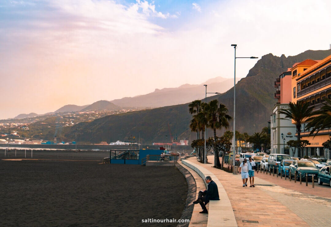 A man sits on a bench by a paved walkway next to a black sand beach in La Palma, Canary Islands, with mountains and buildings in the background under a hazy sky—a perfect scene from any travel guide.