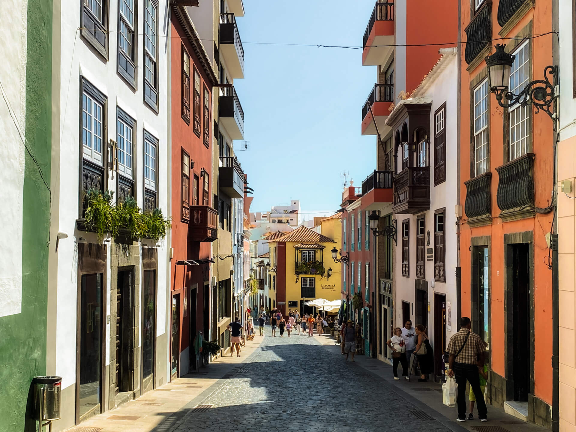 A narrow cobblestone street lined with colorful buildings in La Palma, Canary Islands, welcomes pedestrians strolling along the sidewalks, leading to a bright yellow building at the street’s end under a clear sky—a scene fit for any travel guide.