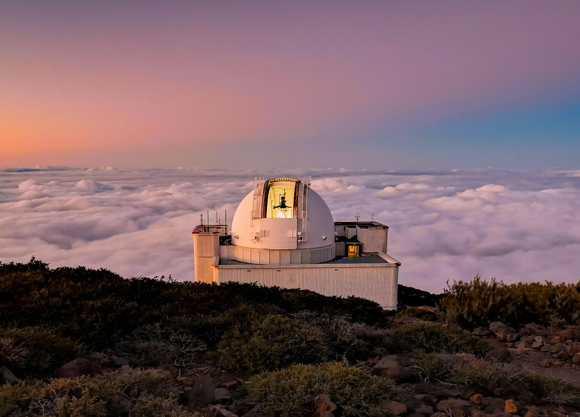 A white observatory dome sits on a mountaintop above a blanket of clouds at sunset on La Palma in the Canary Islands, with vegetation in the foreground and a person visible inside the dome.