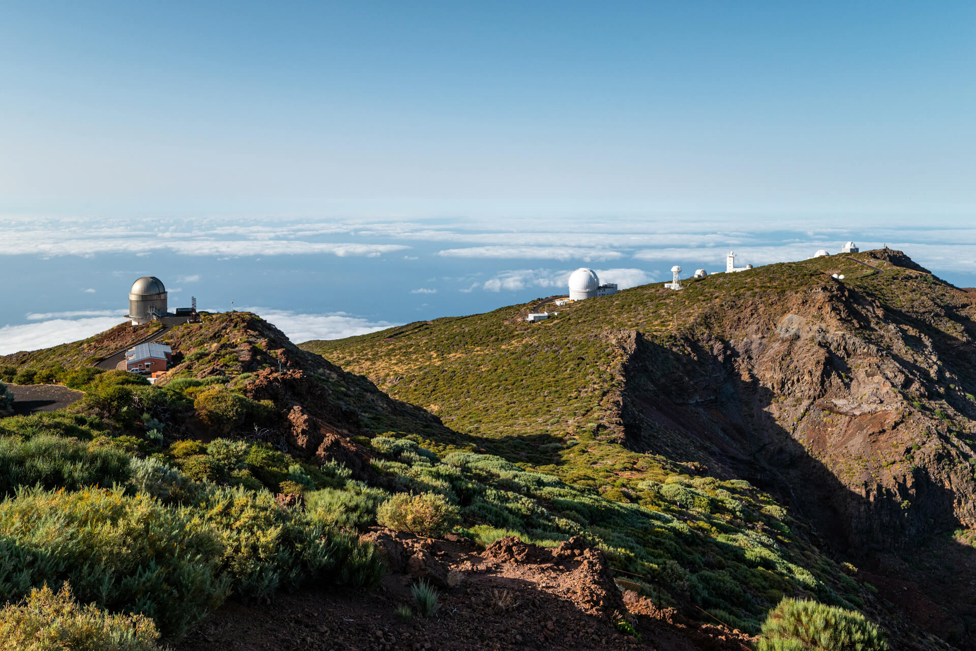 Several observatory domes sit atop a green, rocky mountain ridge under a clear sky on La Palma, one of the Canary Islands, with clouds visible in the distance—a perfect scene for any travel guide.