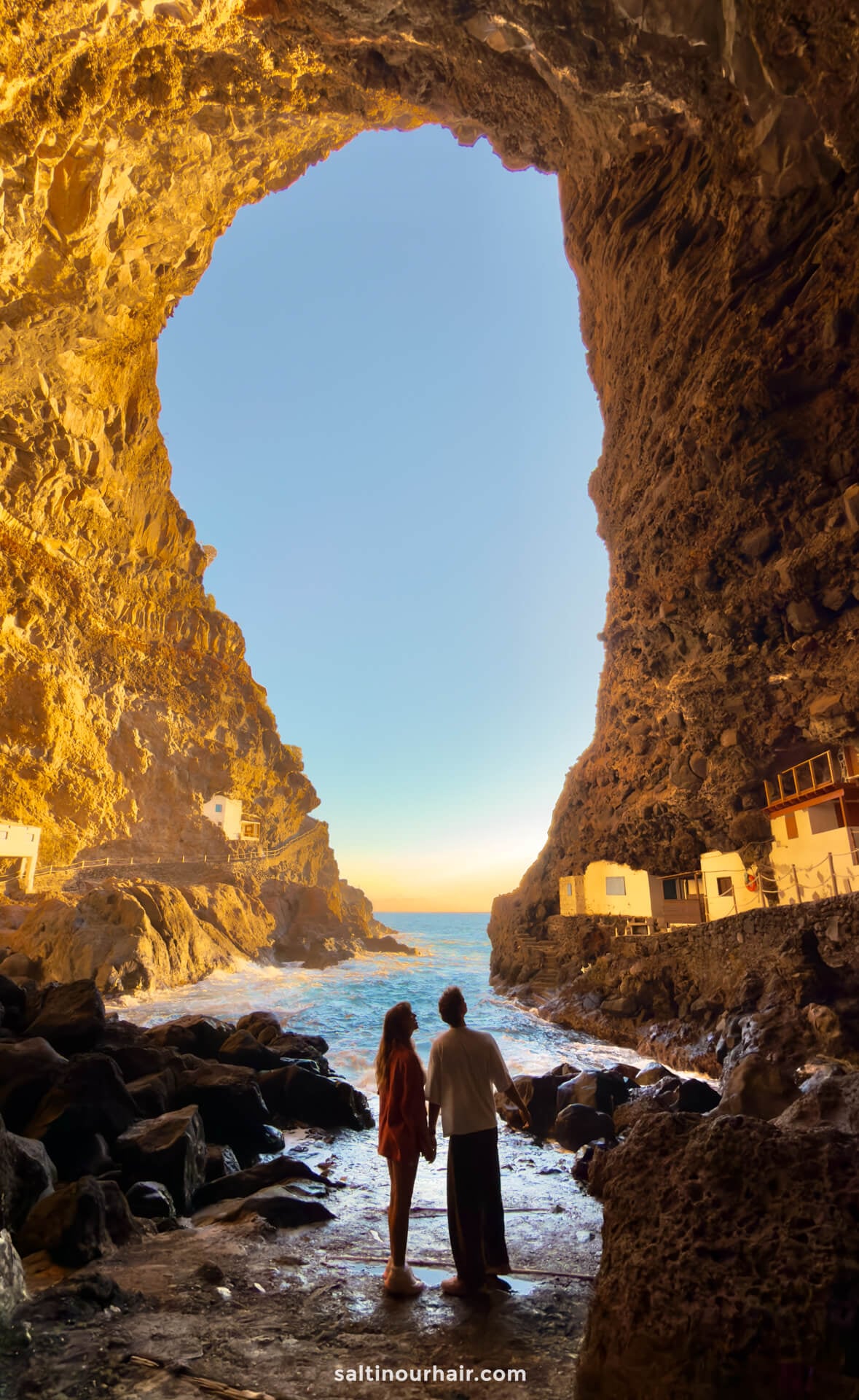 Two people stand inside a large rocky cave on La Palma, holding hands and looking out towards the sea, with sunlight illuminating the cave walls and small buildings nearby—a perfect moment in the Canary Islands for any travel guide.