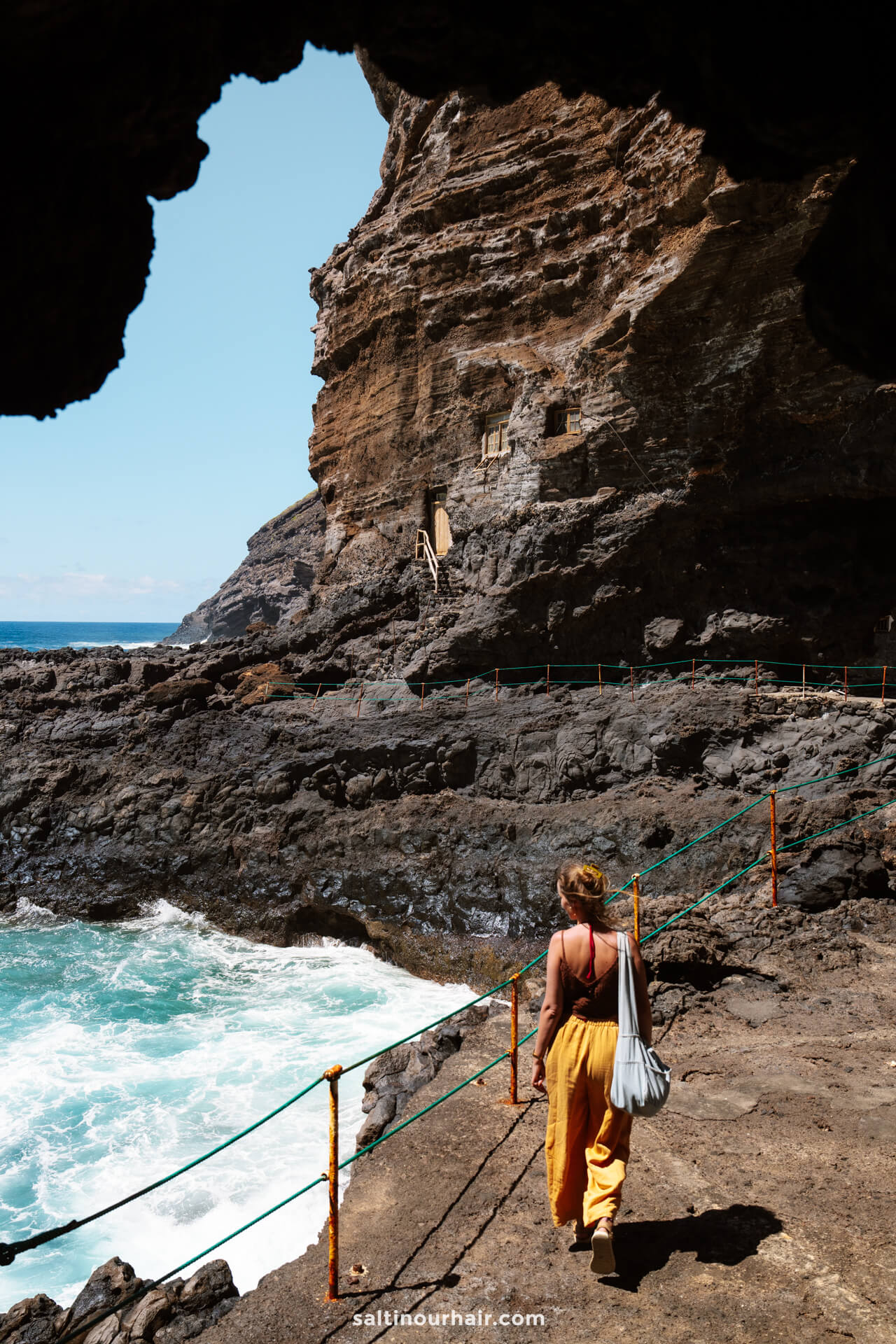 A person walks along a rocky coastal path in La Palma, Canary Islands, with waves crashing below and cliffs rising under a partly cloudy sky—a perfect scene from a travel guide.