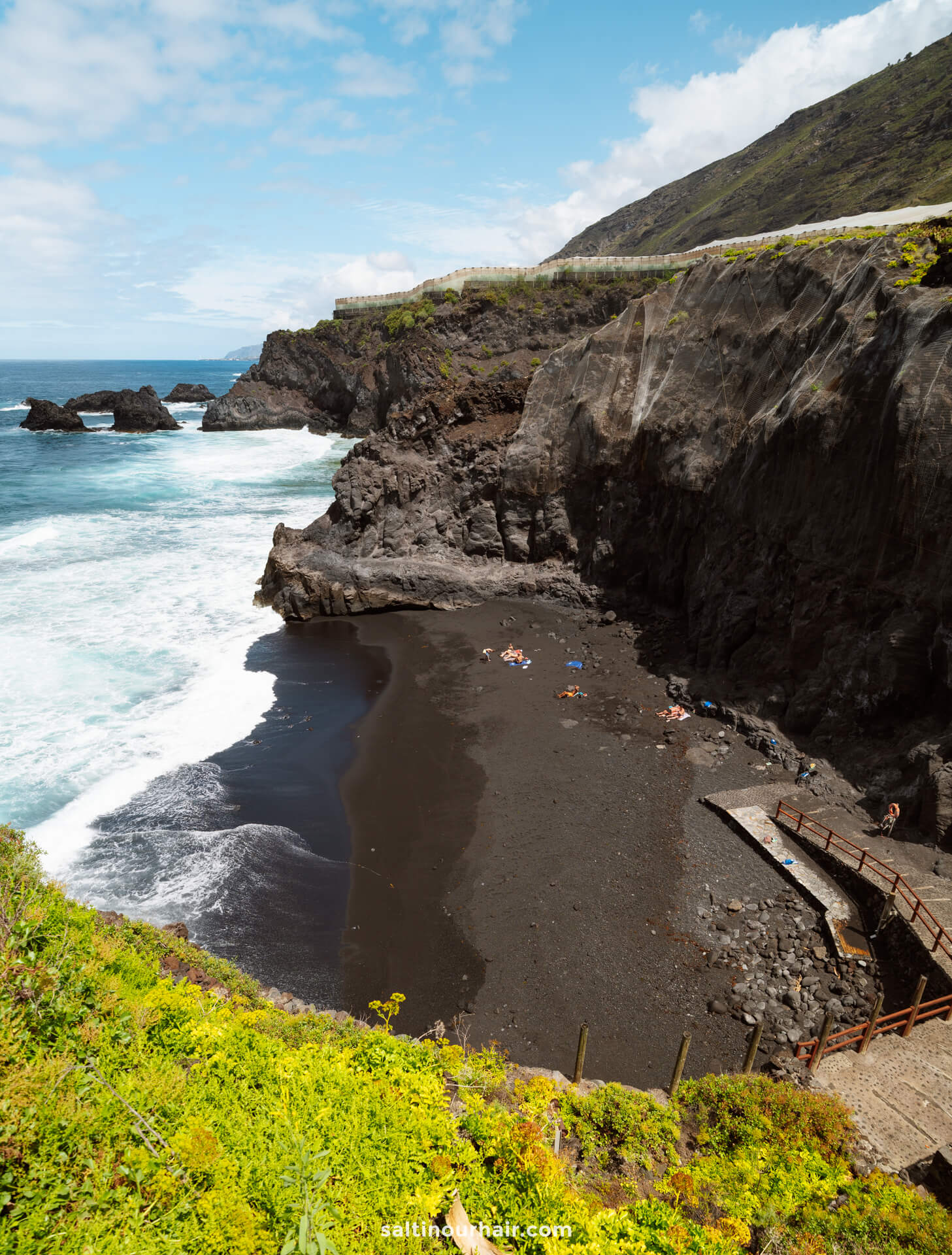 Tucked away on La Palma in the Canary Islands, this small black sand beach is surrounded by high rocky cliffs and waves, with a few people sunbathing and a wooden walkway leading down to the shore—a hidden gem for any travel guide.