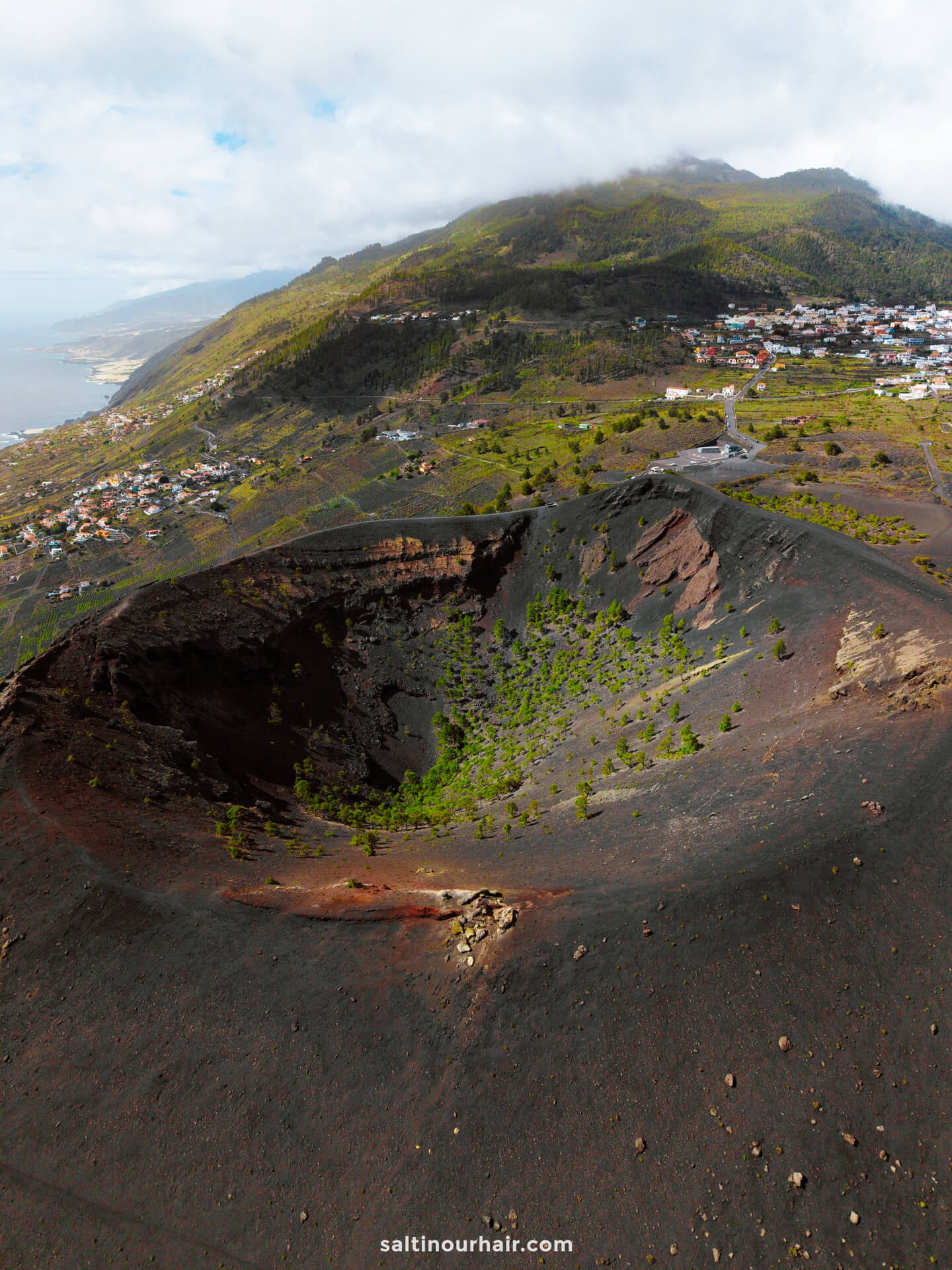 Aerial view of a large volcanic crater with sparse vegetation on La Palma, Canary Islands, surrounded by hills, houses, and a distant coastline under a partly cloudy sky—perfect for any travel guide.