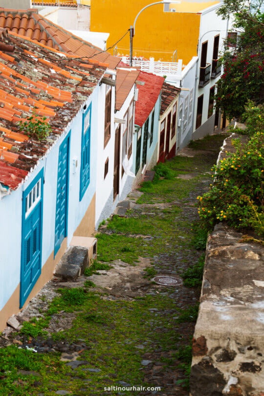 Narrow cobblestone alley lined with colorful houses and blue doors, with greenery on one side and a stone wall in the foreground—perfect for any La Palma travel guide to the Canary Islands.