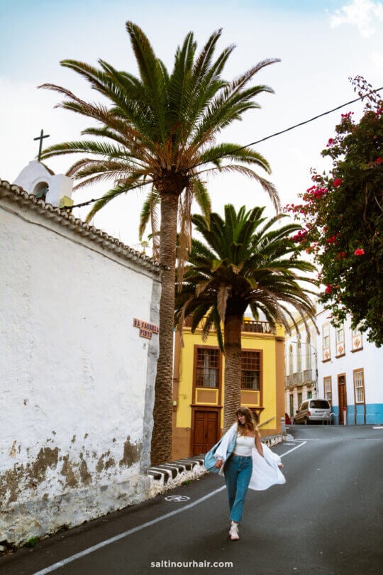 A woman in jeans and a white shirt walks on a narrow street lined with palm trees and colorful buildings in La Palma, Canary Islands, with a church visible on the left—a perfect scene for any Travel Guide.