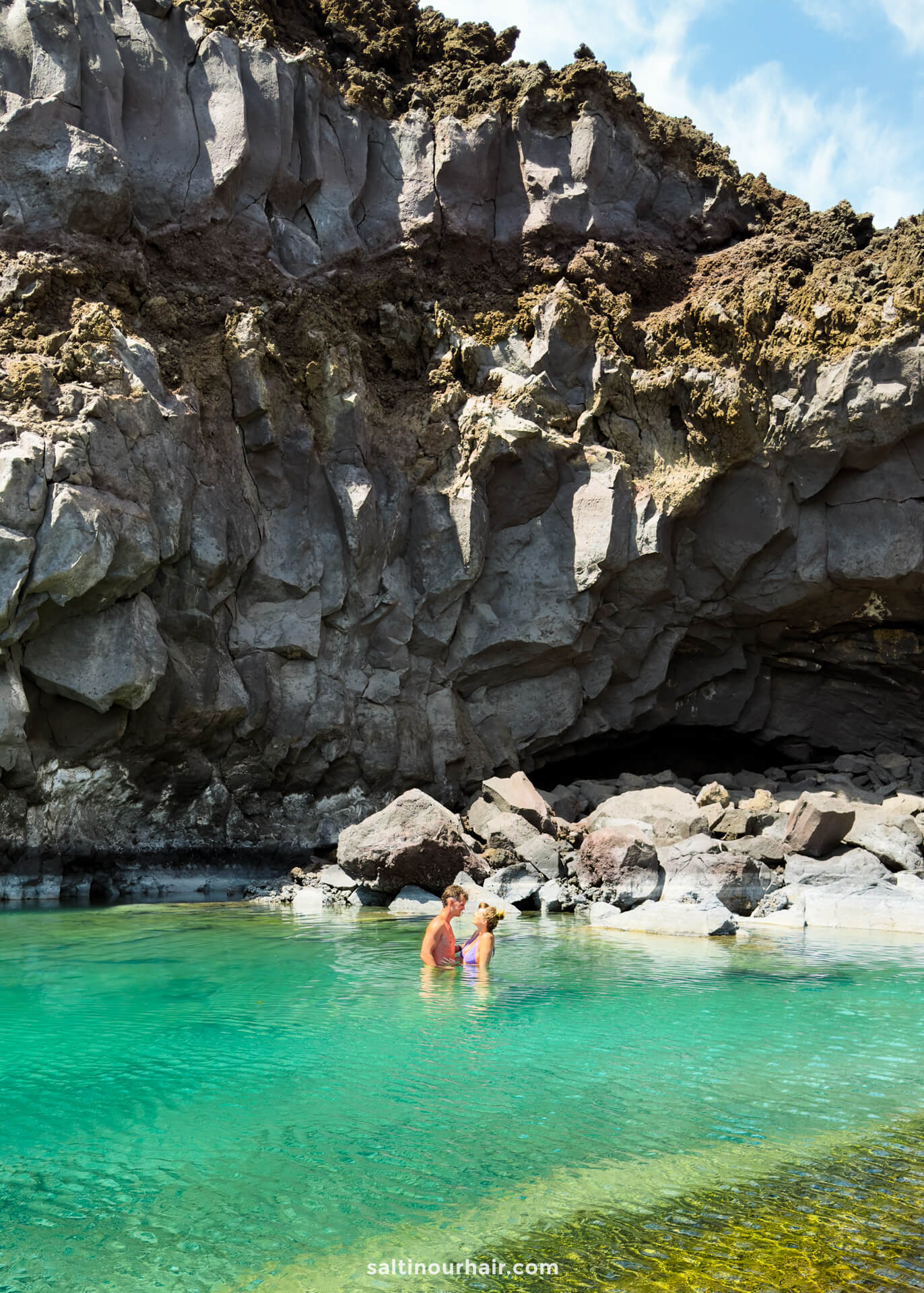 Two people stand in clear turquoise water near rocky cliffs and a small cave under a partly cloudy sky, capturing the natural beauty of La Palma in the Canary Islands—a perfect scene for any travel guide.