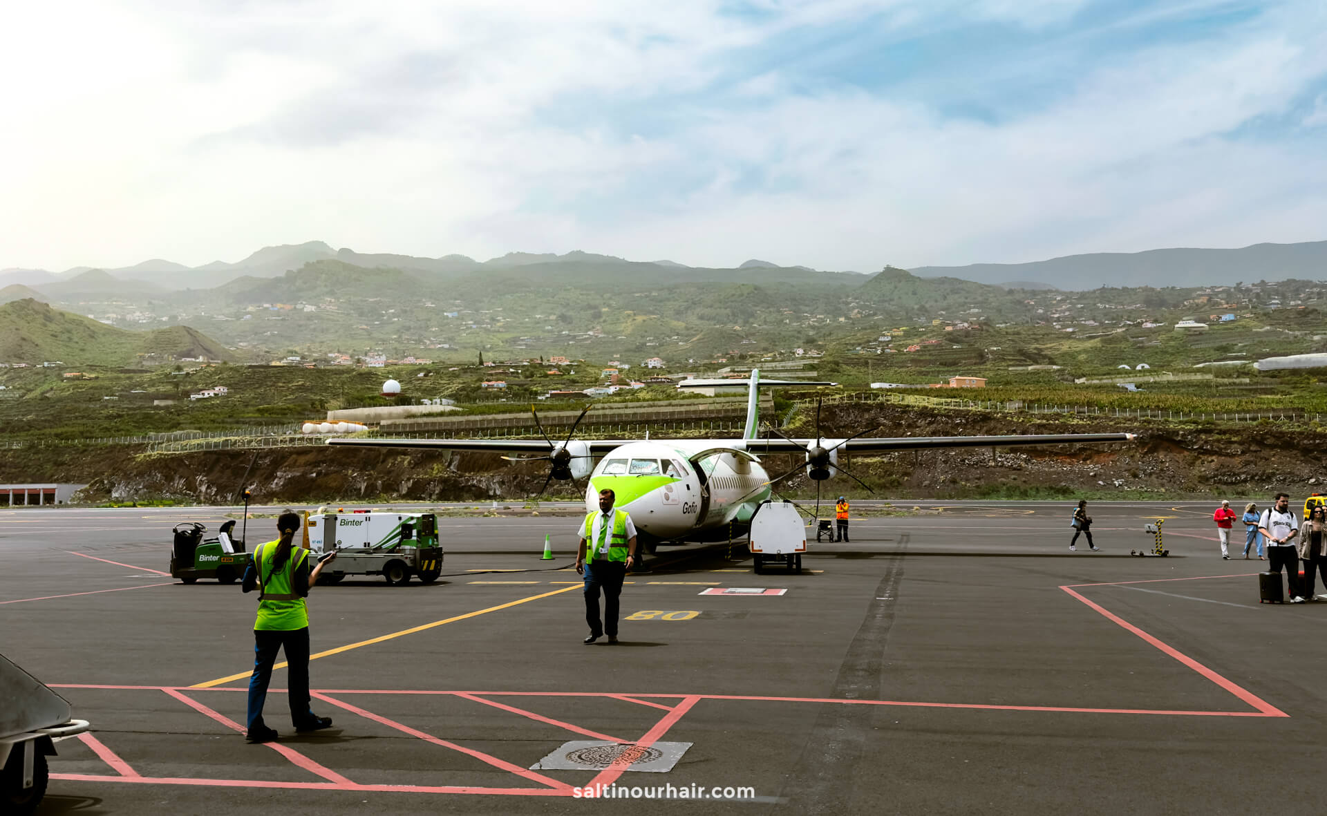A small passenger plane is parked on an airport tarmac in La Palma, with ground crew and equipment nearby; green hills and houses are visible in the background under a partly cloudy Canary Islands sky.