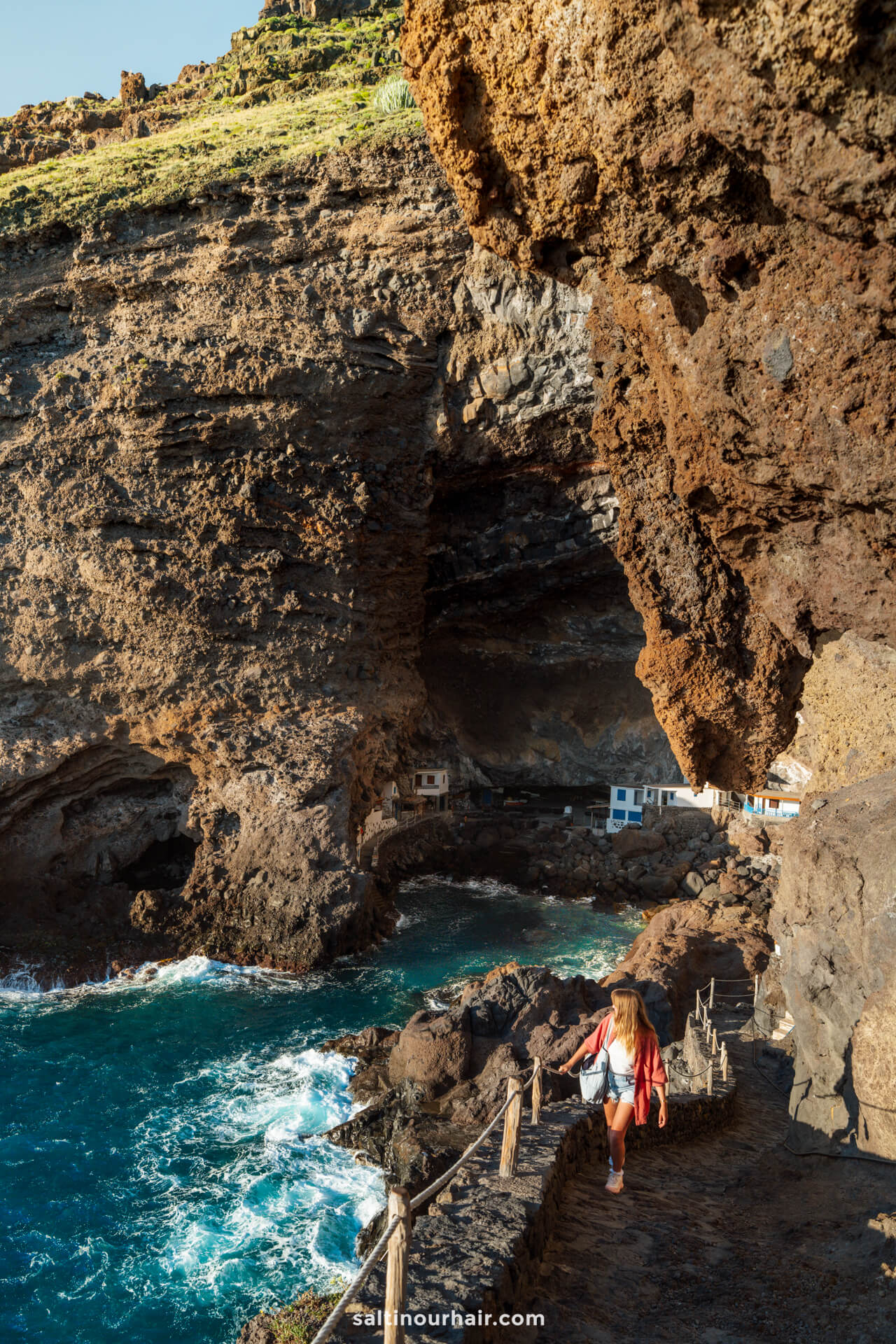 A woman walks along a rocky cliffside path above blue ocean water on La Palma, heading toward small white structures built into the cave-like rock formation in the Canary Islands.