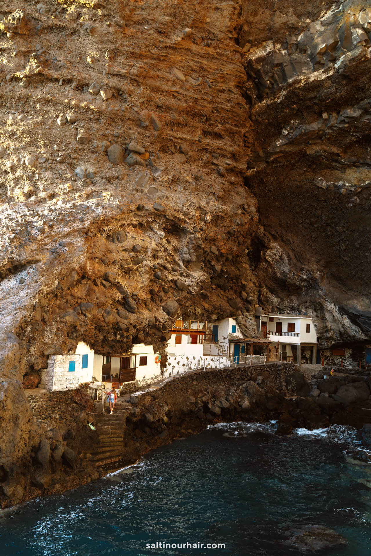 Small white houses built into a large rocky cave overlook blue ocean water on La Palma in the Canary Islands, with stairs leading down to the shore—a hidden gem for any travel guide.