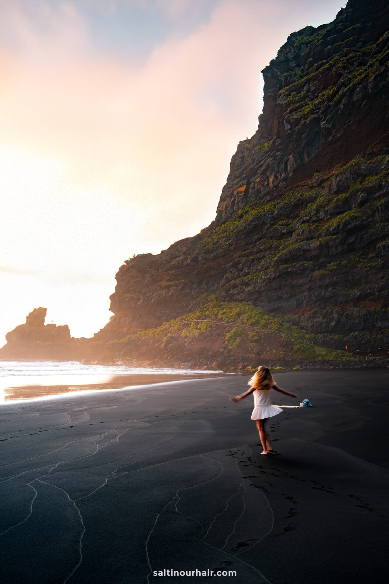A woman in a white dress stands on a black sand beach near tall cliffs at sunset in La Palma, Canary Islands, her footprints trailing behind—an inspiring scene for any travel guide.