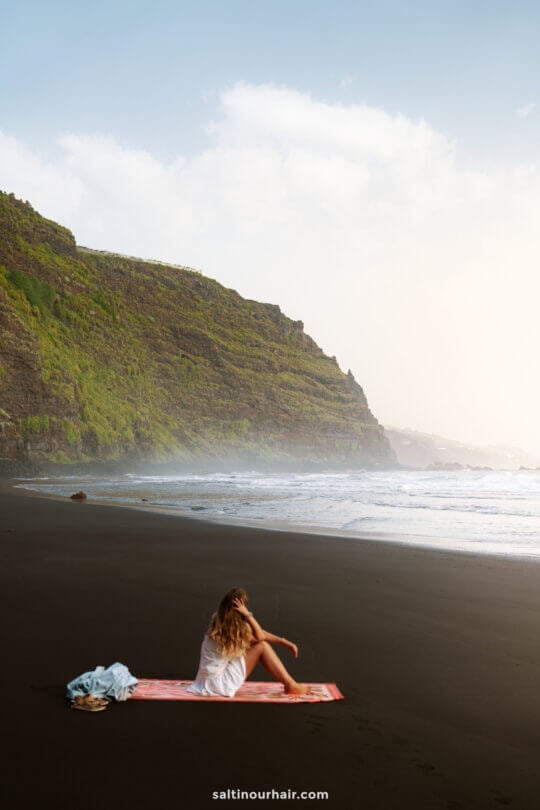 A woman sits on a towel on a black sand beach in La Palma near the ocean, with green cliffs in the background under a clear sky—a perfect moment from any Canary Islands travel guide.