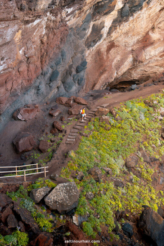 A person walks up a dirt path with wooden steps in a rocky, cliffside area with green vegetation and large boulders on La Palma, one of the scenic Canary Islands.