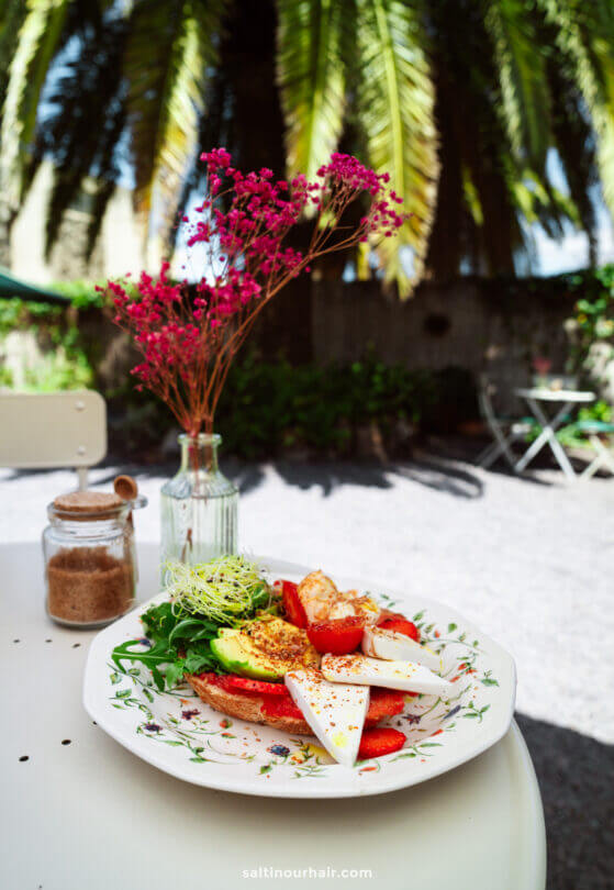 A plate of sliced cheese, tomatoes, avocado, and greens on toast sits on a white table outdoors in La Palma, with a jar of brown sugar and a vase of pink flowers beside it—perfect for a Canary Islands travel guide.
