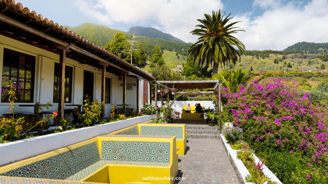 A paved outdoor seating area with yellow-tiled benches, flowering shrubs, and a view of mountains and palm trees under a partly cloudy sky, perfect for any Canary Islands travel guide or inspiration from beautiful La Palma.